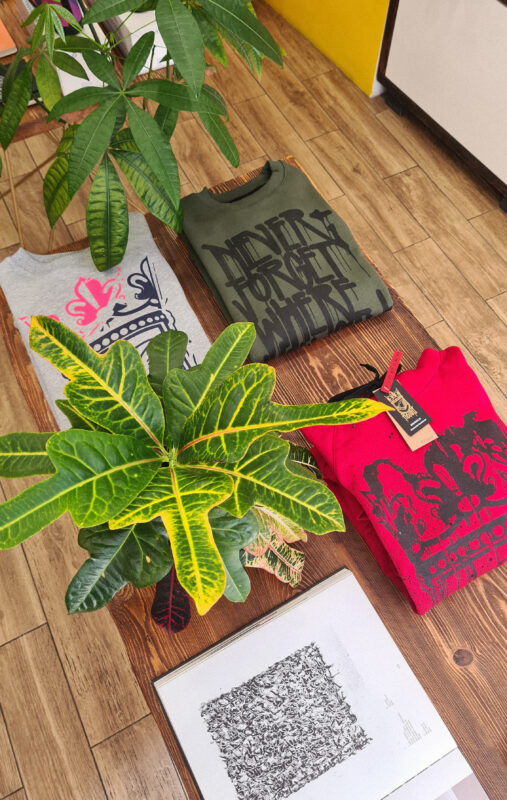 Three folded t-shirts in gray, green, and red with graphic designs, surrounded by a potted plant and a framed artwork.