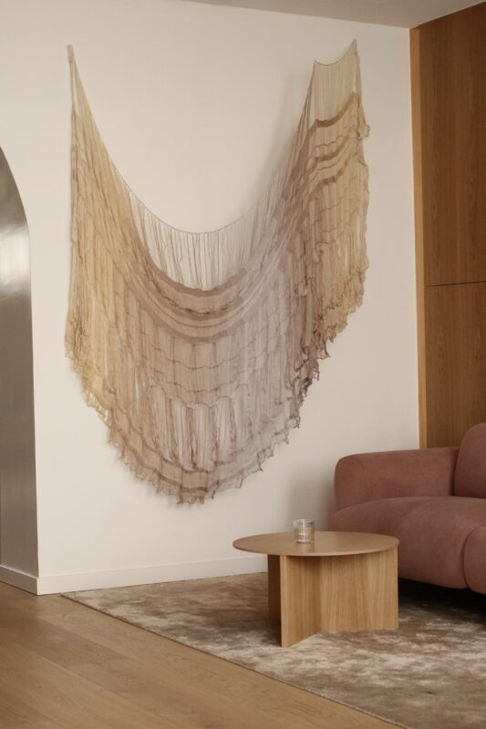 Translucent wall hanging with layered textures in neutral tones, displayed above a pink sofa and wooden coffee table.