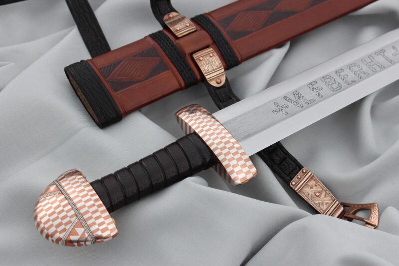 Sword with a checkered hilt and engraved blade, resting on a gray fabric alongside a decorative scabbard.