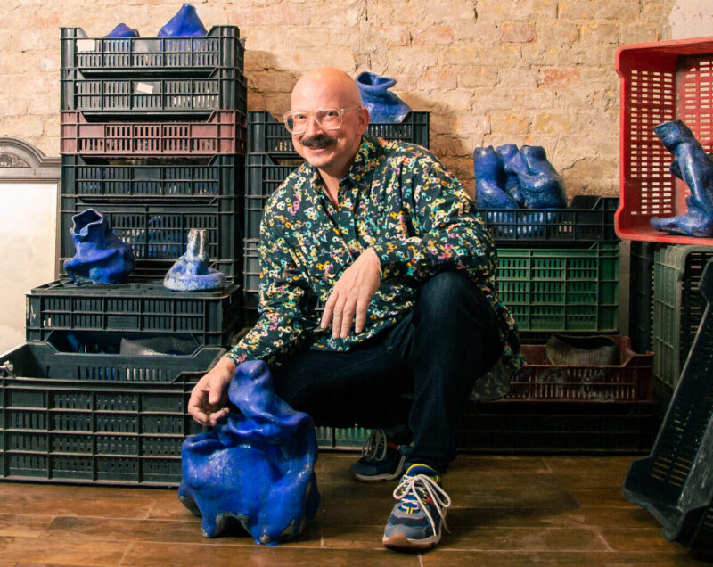 Simon Holpert, wearing a floral shirt, kneels beside blue sculptures in a studio filled with crates.