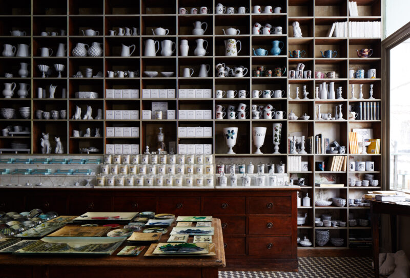 Wooden shelves filled with ceramic items and curiosities, with a display table in front showcasing various artworks.