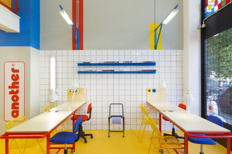 Brightly colored nail studio interior with white tiled walls, yellow floor, and workstations featuring red and blue chairs.