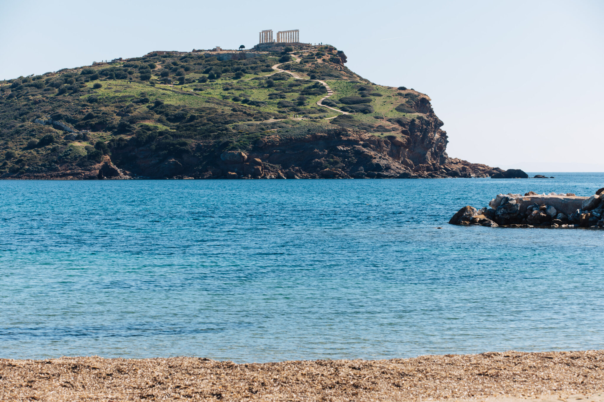 Clear blue sea with a sandy beach in the foreground, and a green hill with ancient ruins in the background.