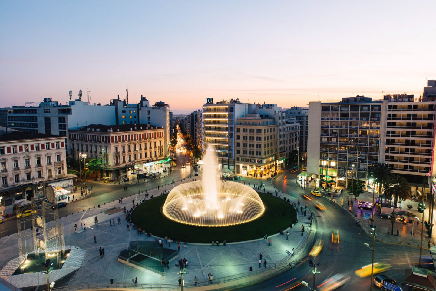 Vibrant night scene in Athens with a fountain illuminated, surrounded by bustling streets and buildings.