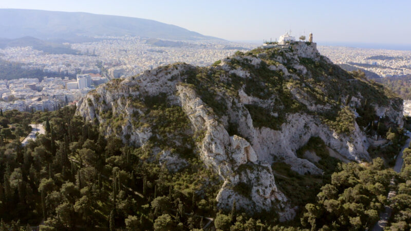 Aerial view of a rocky hill surrounded by greenery, overlooking Athens and the Aegean Sea in the background.