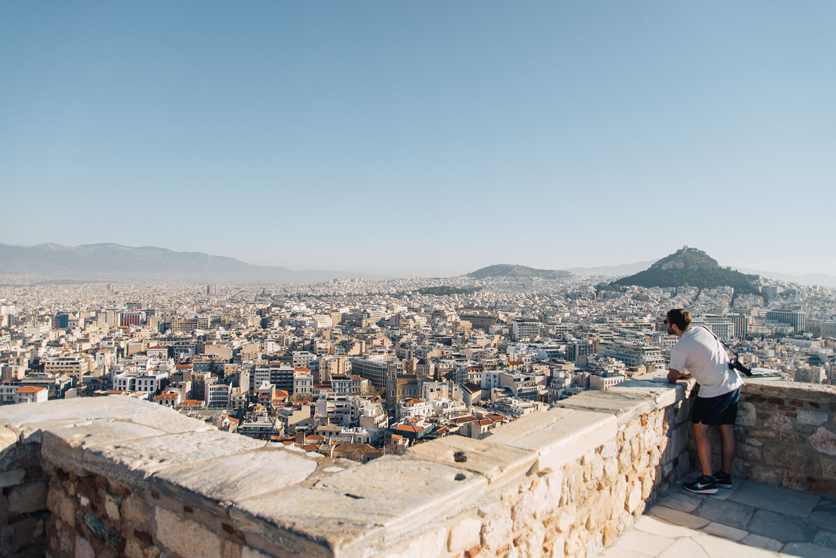 Panoramic view from Acropolis Hill overlooking Athens, with a person leaning on a stone wall.