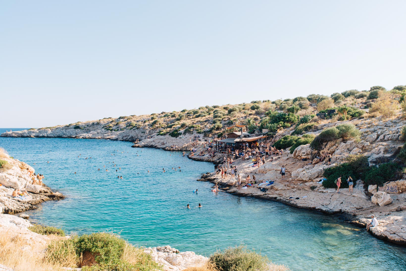 Aegean coastline in Athens Riviera with people swimming and a beach bar amidst rocky shores and greenery.