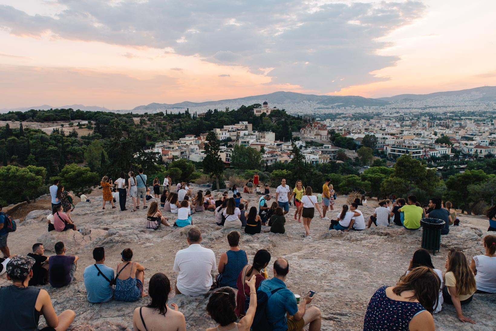 Crowd of people sitting on Areios Pagos Hill at sunset, overlooking Athens and its glowing skyline.