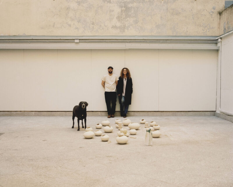 A man and a woman stand beside a dog, surrounded by smooth, light-colored stones in an empty space.