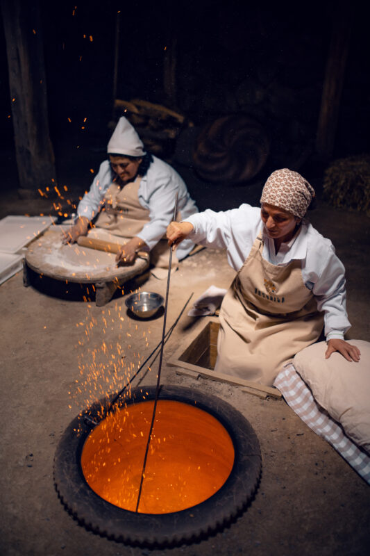 Two women in traditional attire prepare lavash in a dimly lit kitchen, with one stirring a glowing pit while sparks fly.