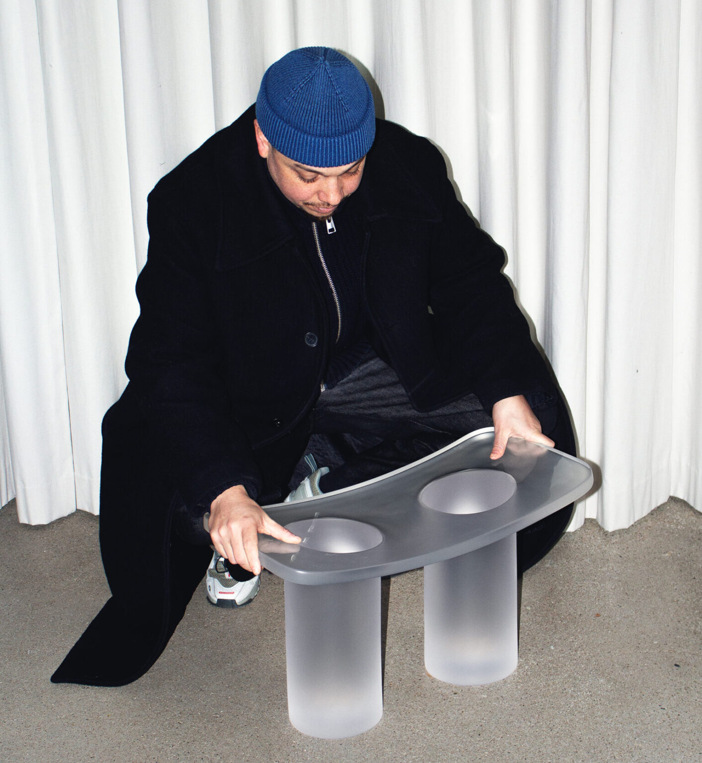Ciro Timmers examines a translucent furniture piece with two circular openings, wearing a blue beanie and black coat.