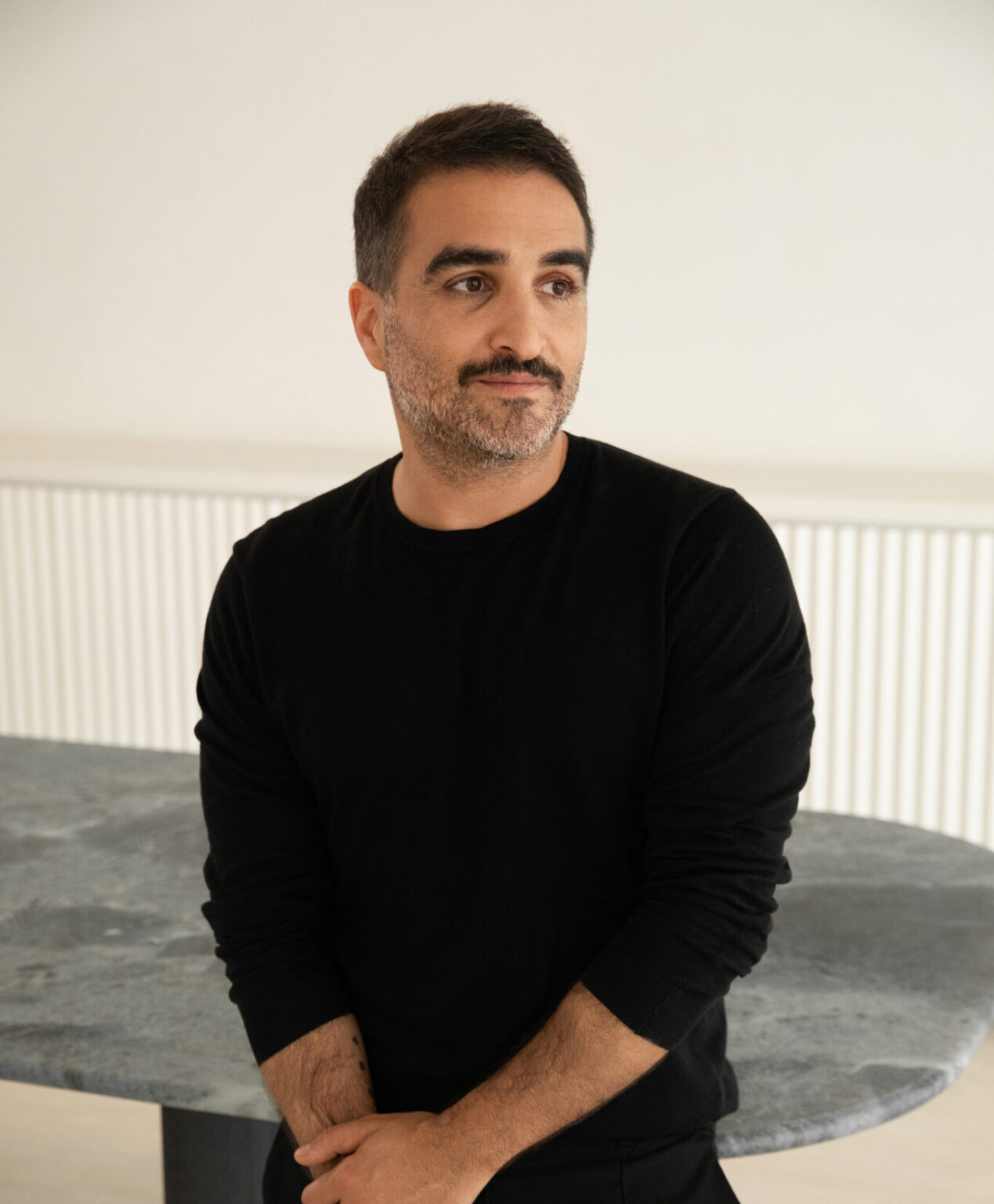 A man with short dark hair and a beard stands in a black sweater, looking thoughtfully to the side, in a minimalist interior.