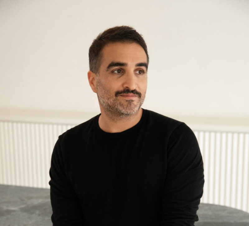 A man with short dark hair and a beard stands in a black sweater, looking thoughtfully to the side, in a minimalist interior.