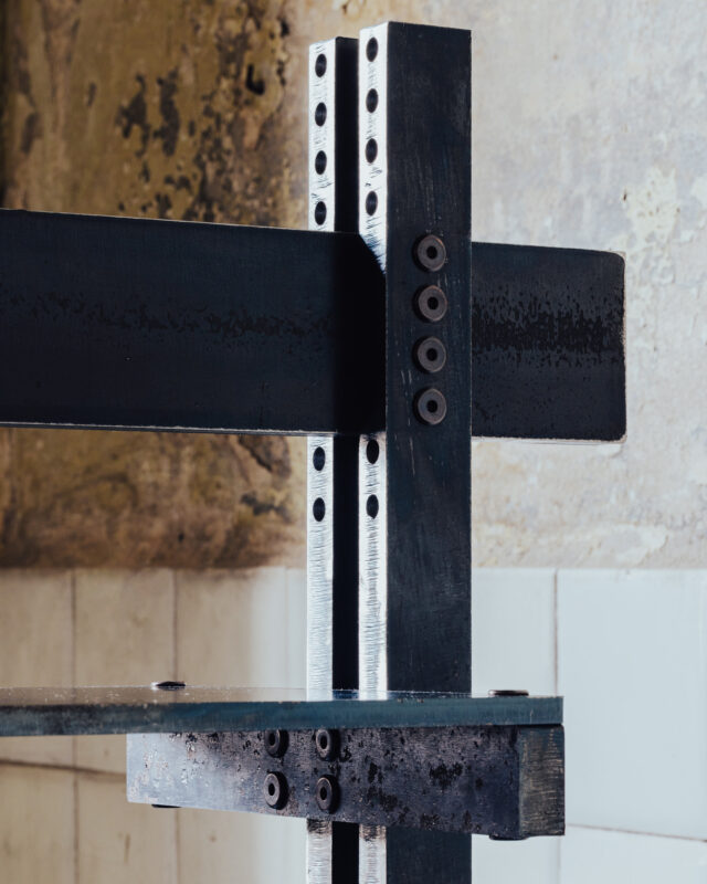 Close-up of a metallic structure with black beams and bolts, set against a textured wall.