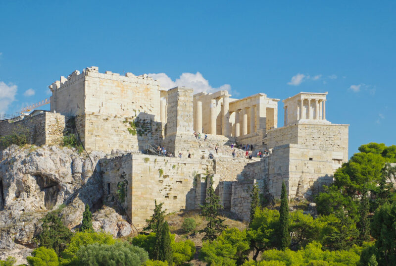 Acropolis of Athens with ancient ruins atop a rocky hill, surrounded by greenery and a clear blue sky.