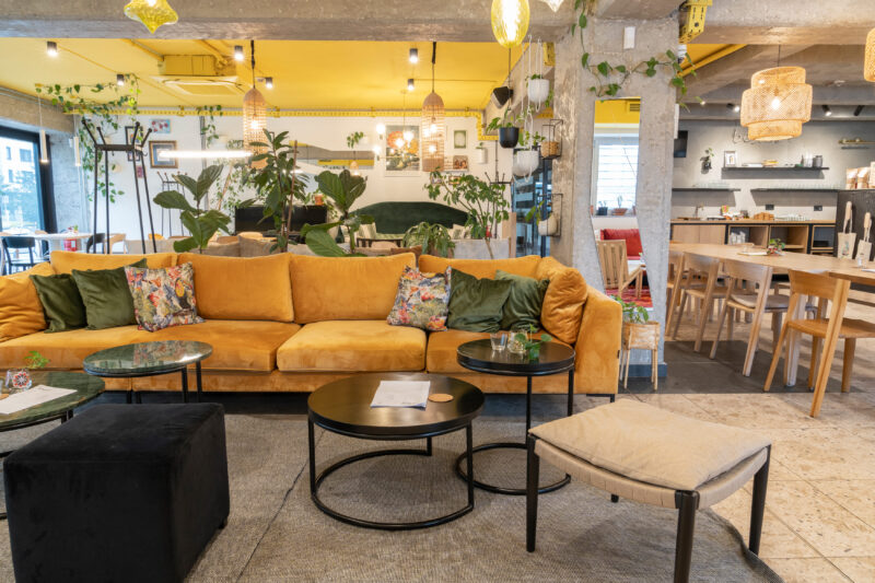 Cozy interior of Bistro Zdravo featuring a yellow sofa with decorative pillows, plants, and wooden dining tables.