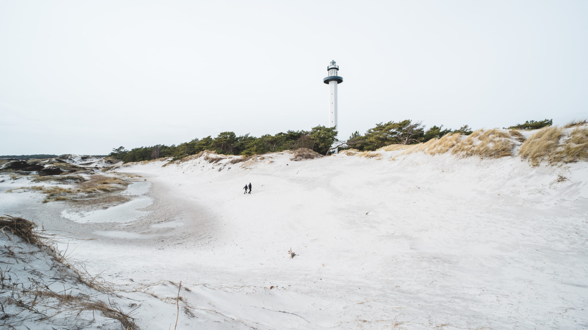 Winter light at Dueodde, featuring a lighthouse above white sand dunes and two figures walking in the distance.