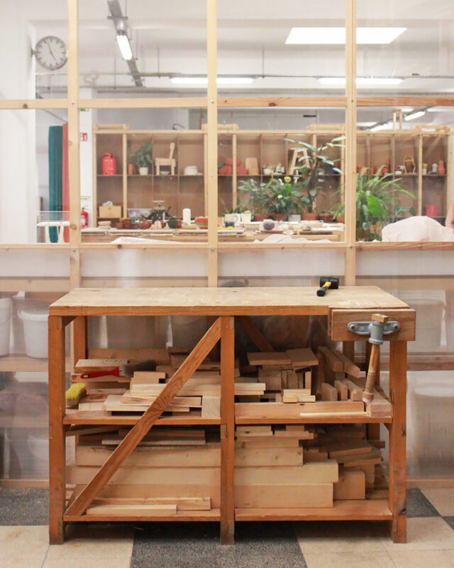 Woodworking workshop with a wooden workbench holding various pieces of lumber, surrounded by shelves of tools and plants.