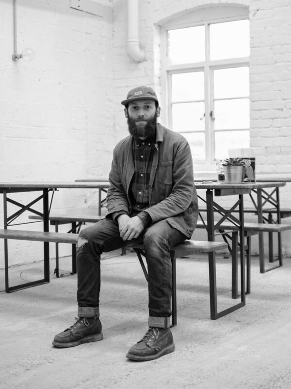 Black and white headshot of Matthew Moore sitting on a bench in a bright, minimalist room with a window.