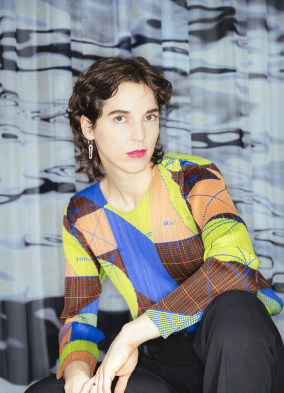 Portrait of a woman with curly hair, wearing a colorful patterned top, seated against a patterned backdrop.