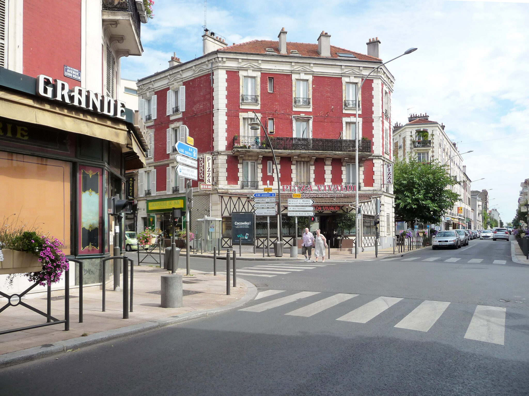 Street view of Le Perreux-sur-Marne featuring a red building, shops, and pedestrians crossing at a zebra crossing.