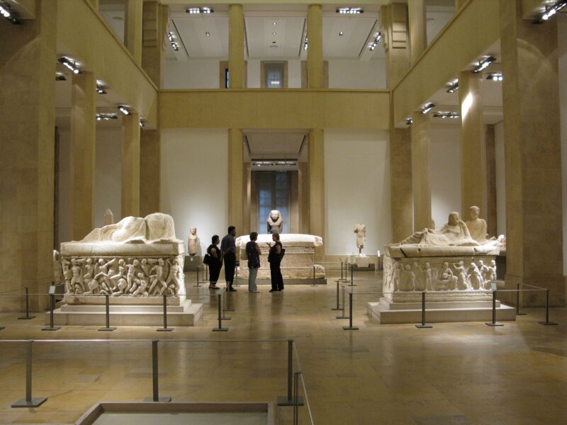 Interior of the National Museum of Beirut, showcasing ancient sculptures and sarcophagi with visitors in the foreground.