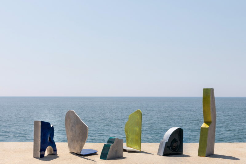 Colorful cast aluminum prototypes arranged on a ledge by the sea, with a clear blue sky and ocean in the background.