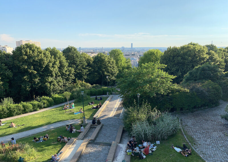 View of Belleville Park in Paris, featuring trees, steps, and people relaxing on the grass under a blue sky.