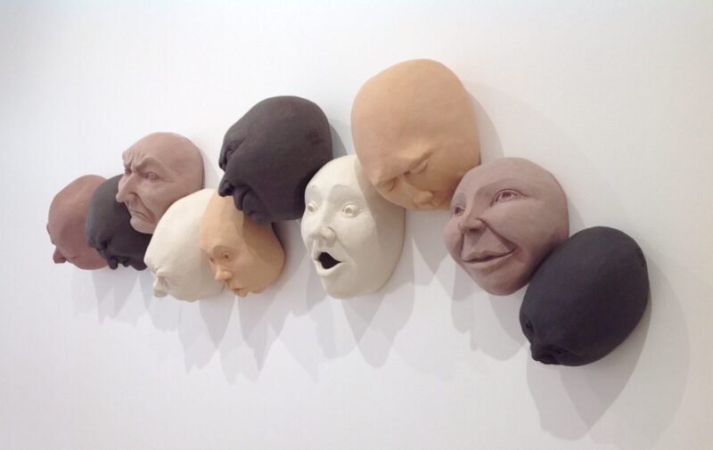 Sculptures of diverse human faces in various skin tones, mounted on a white wall, by artist Sandra Borges.
