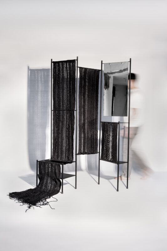 Overall view of a black textile installation with a mirror, featuring a blurred figure walking past.