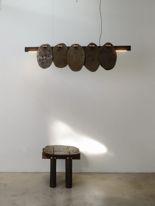 Wooden tabouret with two legs beside five ceramic plates hanging on a white wall, illuminated from above.