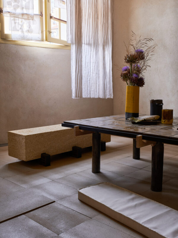 Interior scene featuring a wooden table with ceramics, a textured bench, and a vase of flowers, illuminated by natural light.