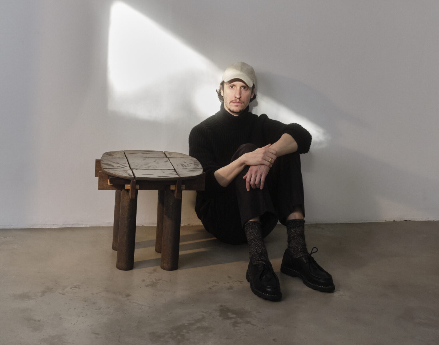Thomas Morineau Barthelemy seated on the floor beside a wooden table, in a well-lit studio setting.