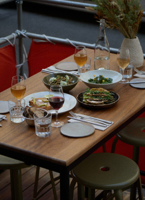 A wooden table set with various dishes, glasses of wine, and water, surrounded by green stools, showcasing a blend of Japa...