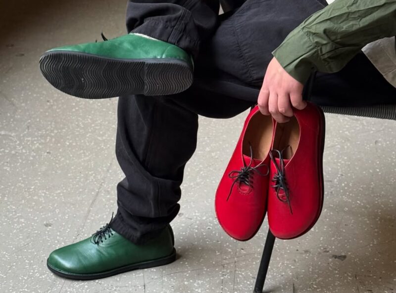 Green and red shoes held by a person sitting on a chair, wearing black pants and a green shirt, in a minimalist setting.