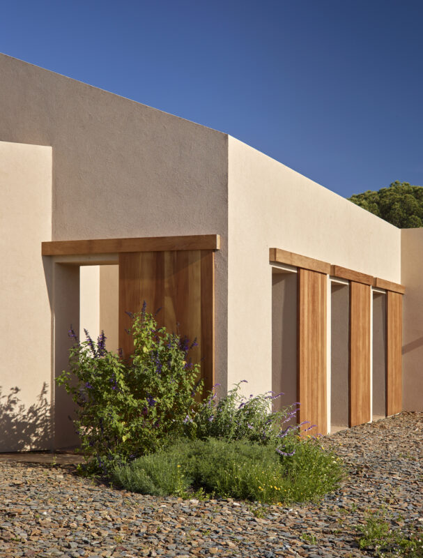 Modern architectural structure with wooden doors, surrounded by greenery and gravel, under a clear blue sky.