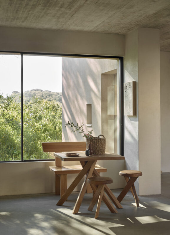 Modern wooden table and stools in a bright room with large window overlooking greenery.