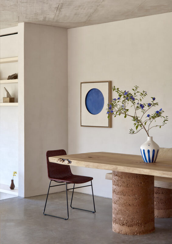 A minimalist dining area featuring a wooden table with a round blue artwork, a vase with flowers, and a red chair.