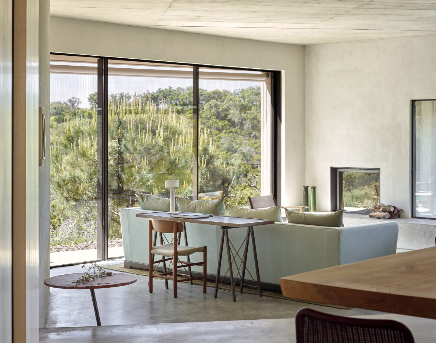 Modern interior of a living space with a light blue sofa, wooden table, and large windows showcasing greenery outside.