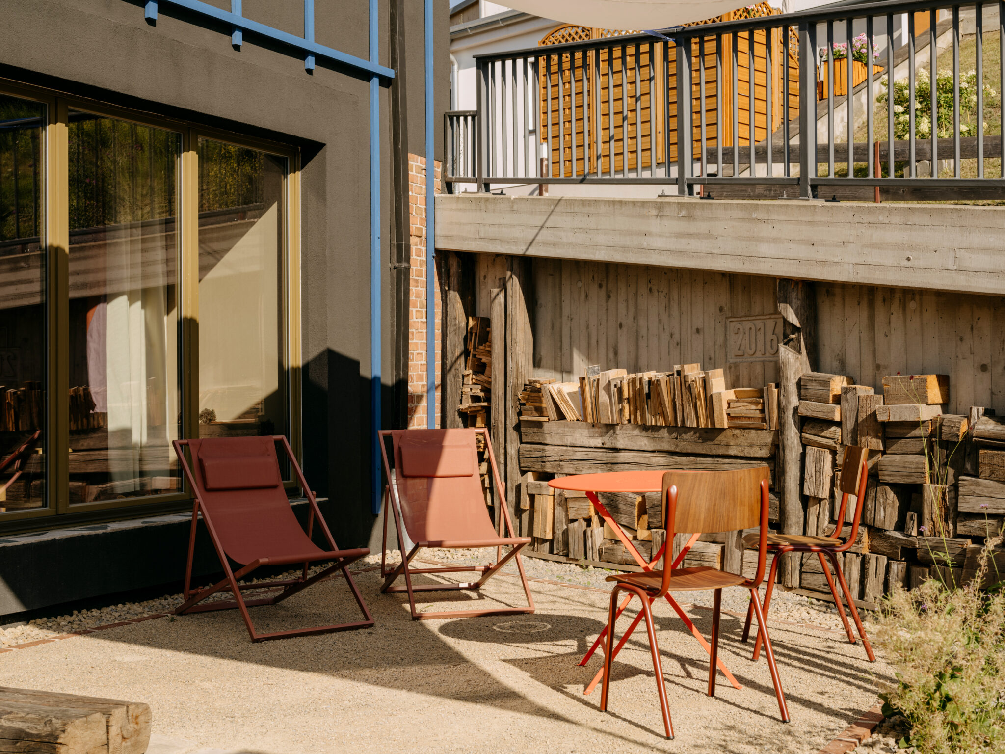 Sunlit patio with two red chairs and a small table in front of a holiday home, surrounded by stacked firewood.