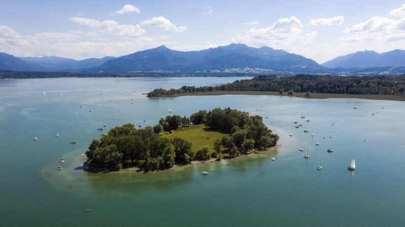 Aerial view of Krautinsel, a small island with greenery, surrounded by water and boats, against a mountainous backdrop.