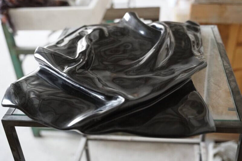 Black, wavy sculpture resting on a glass surface, showcasing smooth, reflective surfaces and intricate textures.