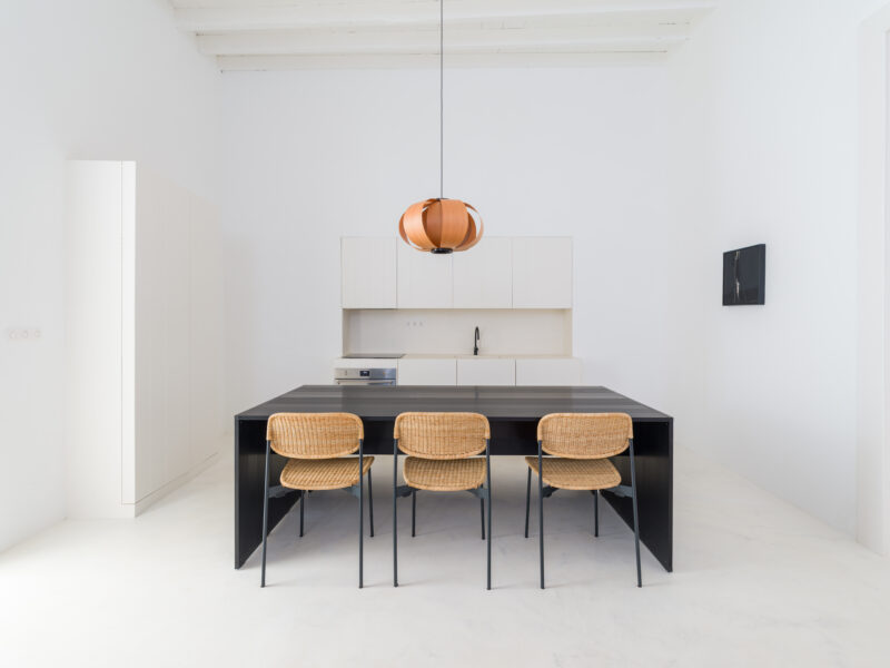 Minimalist kitchen with a black table and four rattan chairs, featuring a pendant light and white walls.