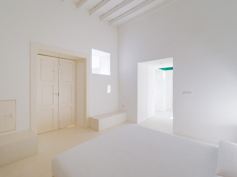 Bright, minimalist bedroom with white walls, a bed, and two doorways leading to other spaces, emphasizing tranquility.