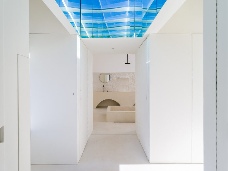 Bright, minimalist interior with white walls, a curved archway, and a blue glass ceiling reflecting light.