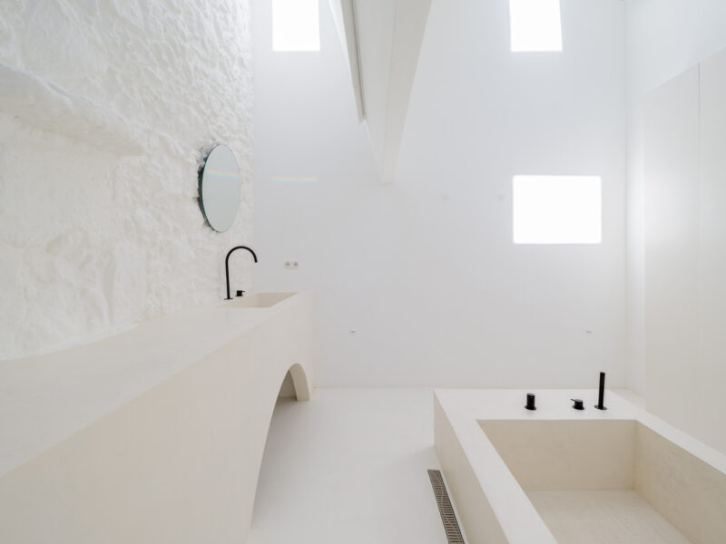 Minimalist bathroom with a stone wall, a round mirror, and a soaking tub, illuminated by bright white light.