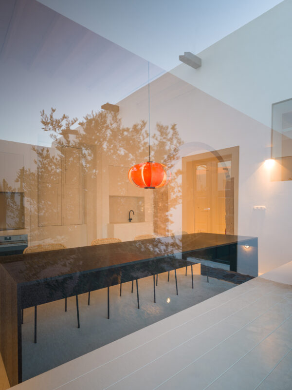 Large open windows connect an interior dining area with a pool, featuring a red pendant light above a dark table.