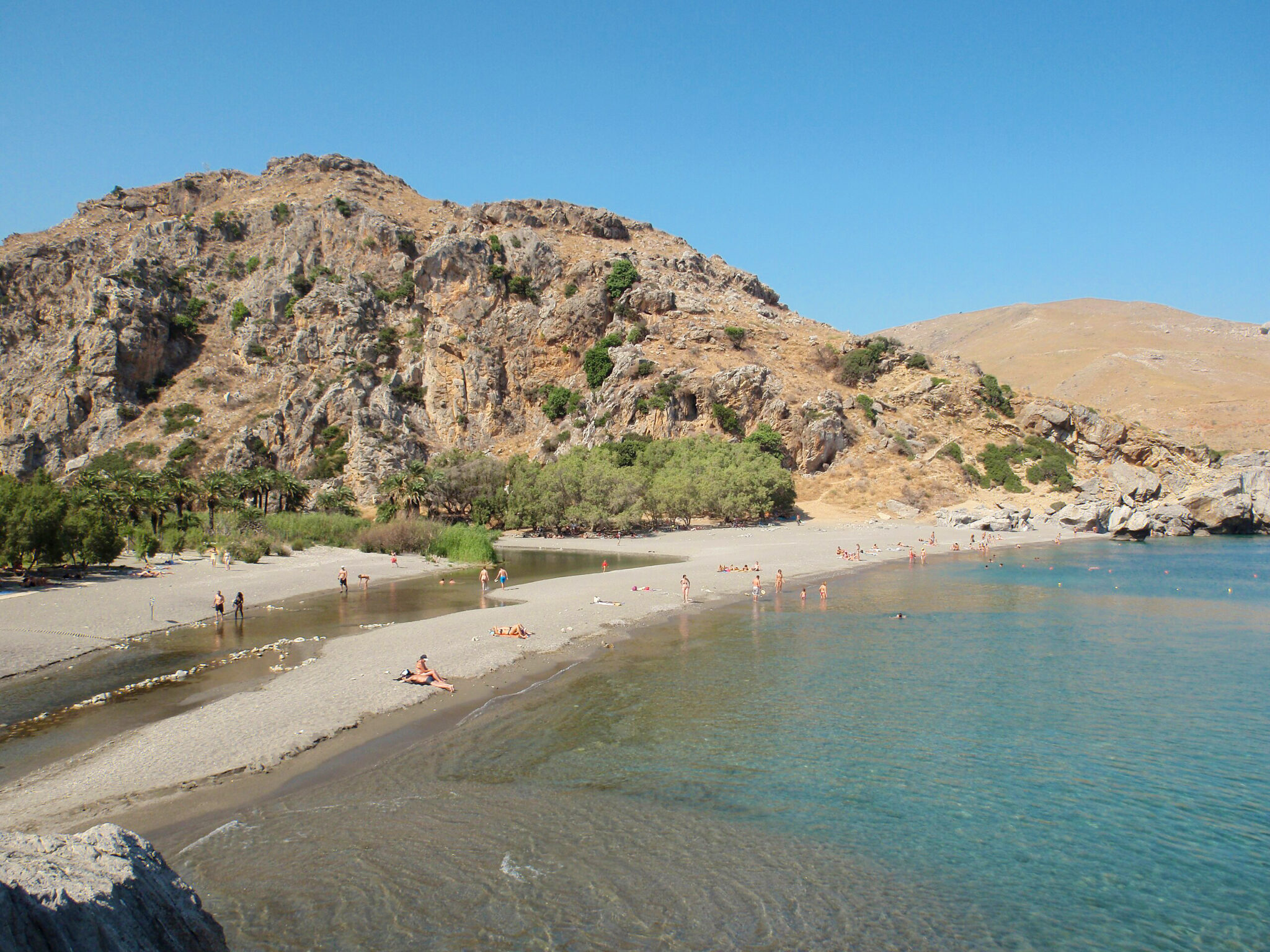 Rethymno beach at Preveli Gorge, surrounded by mountains, with people sunbathing and clear blue water.