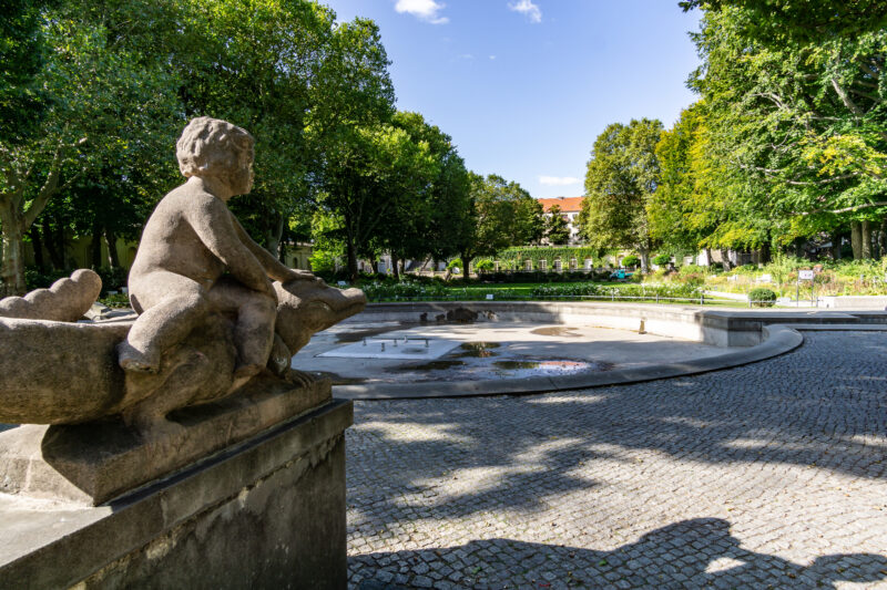 Statue of a cherub sitting on a stone basin at Körnerpark, surrounded by trees and a circular fountain in Berlin-Neukölln.
