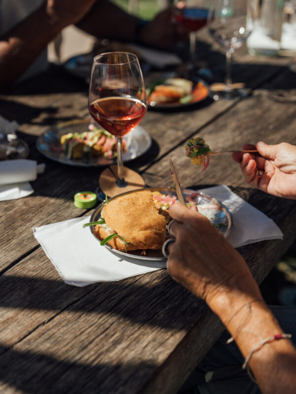 A hand holding a fork and knife, preparing to cut into a sandwich on a plate, with a glass of rosé wine nearby.